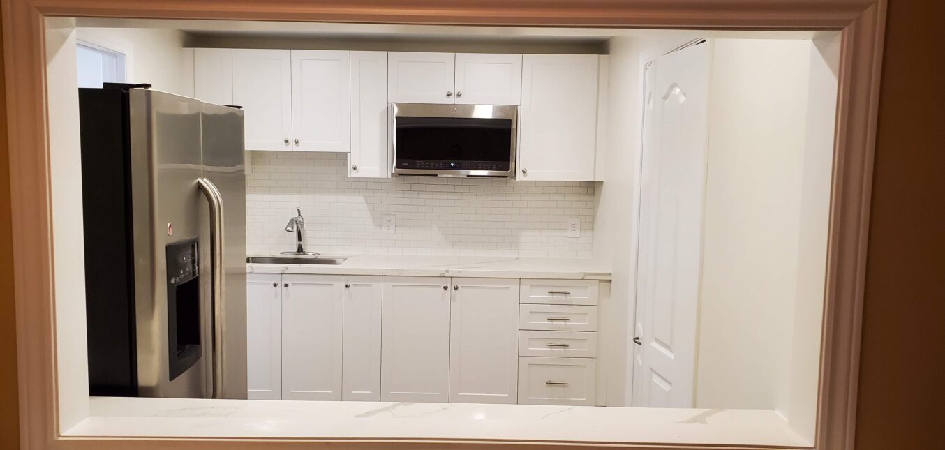 A renovated basement kitchenette with shelves, sink, fridge, and microwave - viewed from a wooden frame window.