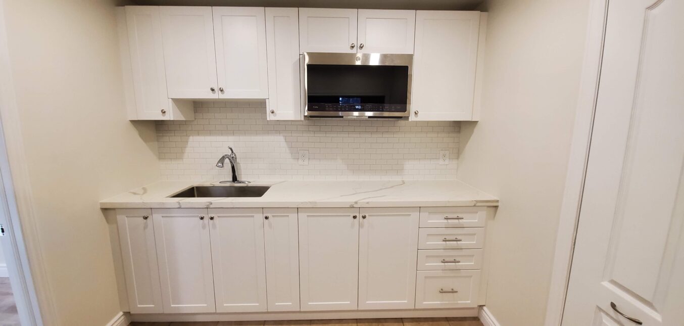 A renovated basement kitchenette with shelves, sink, fridge, and microwave.